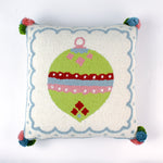 Square hooked wool christmas pillow featuring a green Christmas ornament with red, pink, and blue patterns on a white background with a light blue scalloped border and colorful pom-poms on each corner.