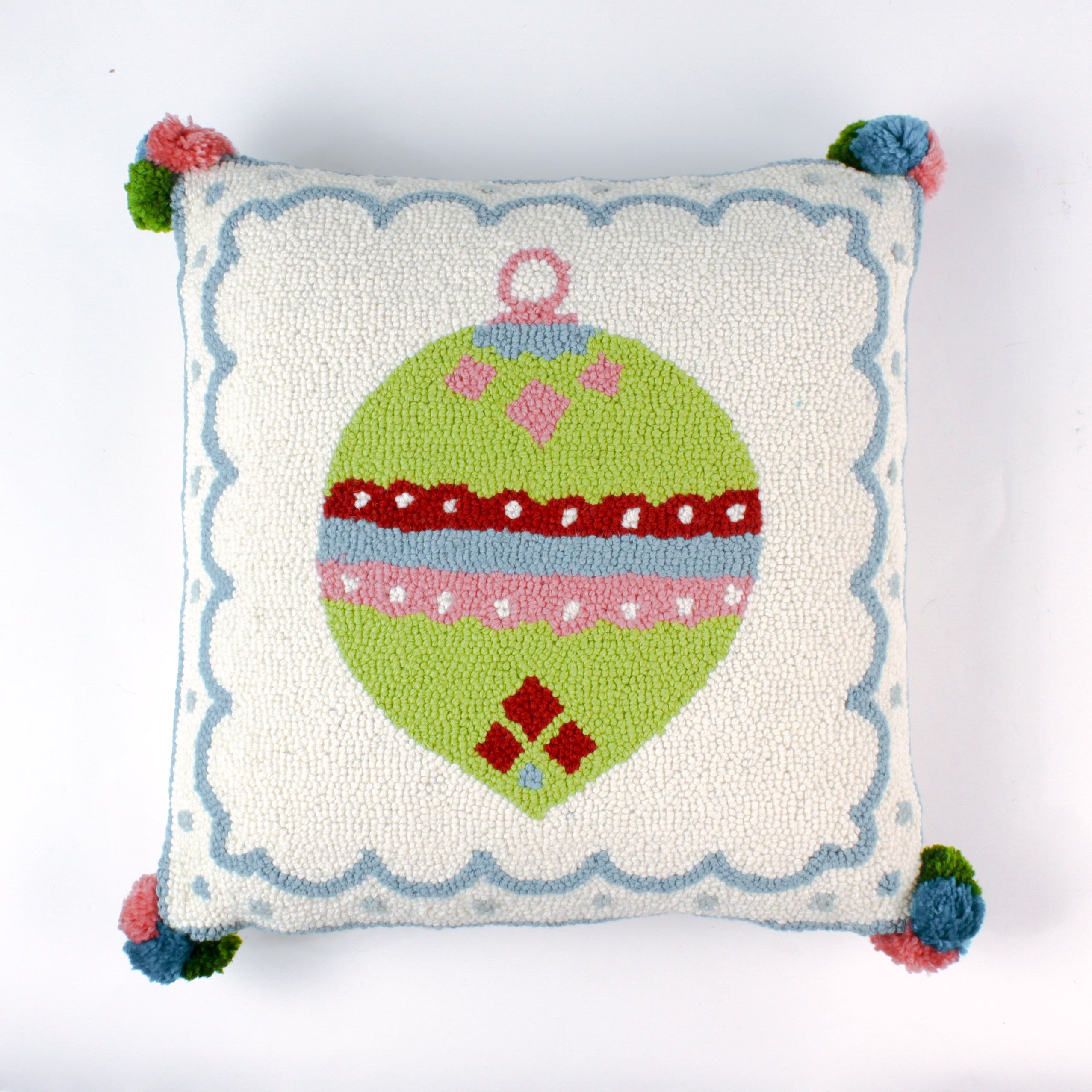 Square hooked wool christmas pillow featuring a green Christmas ornament with red, pink, and blue patterns on a white background with a light blue scalloped border and colorful pom-poms on each corner.