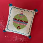 Square hooked wool christmas pillow featuring a green Christmas ornament with red, pink, and blue patterns on a white background with a light blue scalloped border and colorful pom-poms on each corner lying on a red surface