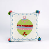 Square hooked wool christmas pillow featuring a green Christmas ornament with red, pink, and blue patterns on a white background with a light blue scalloped border and colorful pom-poms on each corner.