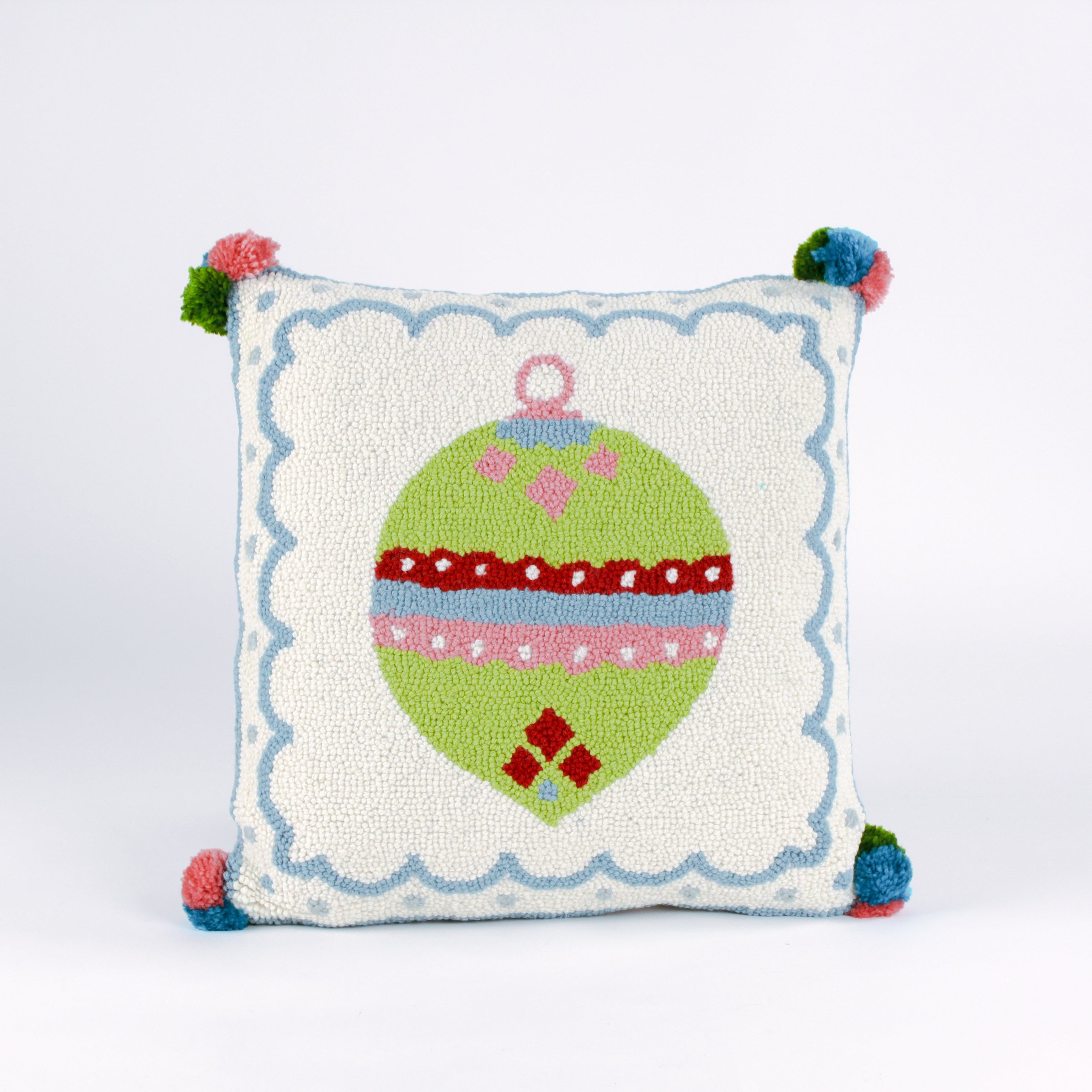 Square hooked wool christmas pillow featuring a green Christmas ornament with red, pink, and blue patterns on a white background with a light blue scalloped border and colorful pom-poms on each corner.