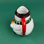 Close-up of a ceramic snowman-shaped pitcher with a red handle, green scarf, and yellow gift detail on a green background