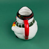 Close-up of a ceramic snowman-shaped pitcher with a red handle, green scarf, and yellow gift detail on a green background