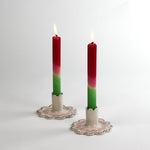 Two lit red-pink-green gradient taper candles in scalloped ceramic candle holders with pink and green detailing, displayed on a white background.