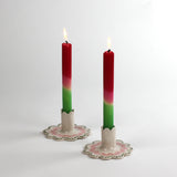 Two lit red-pink-green gradient taper candles in scalloped ceramic candle holders with pink and green detailing, displayed on a white background.