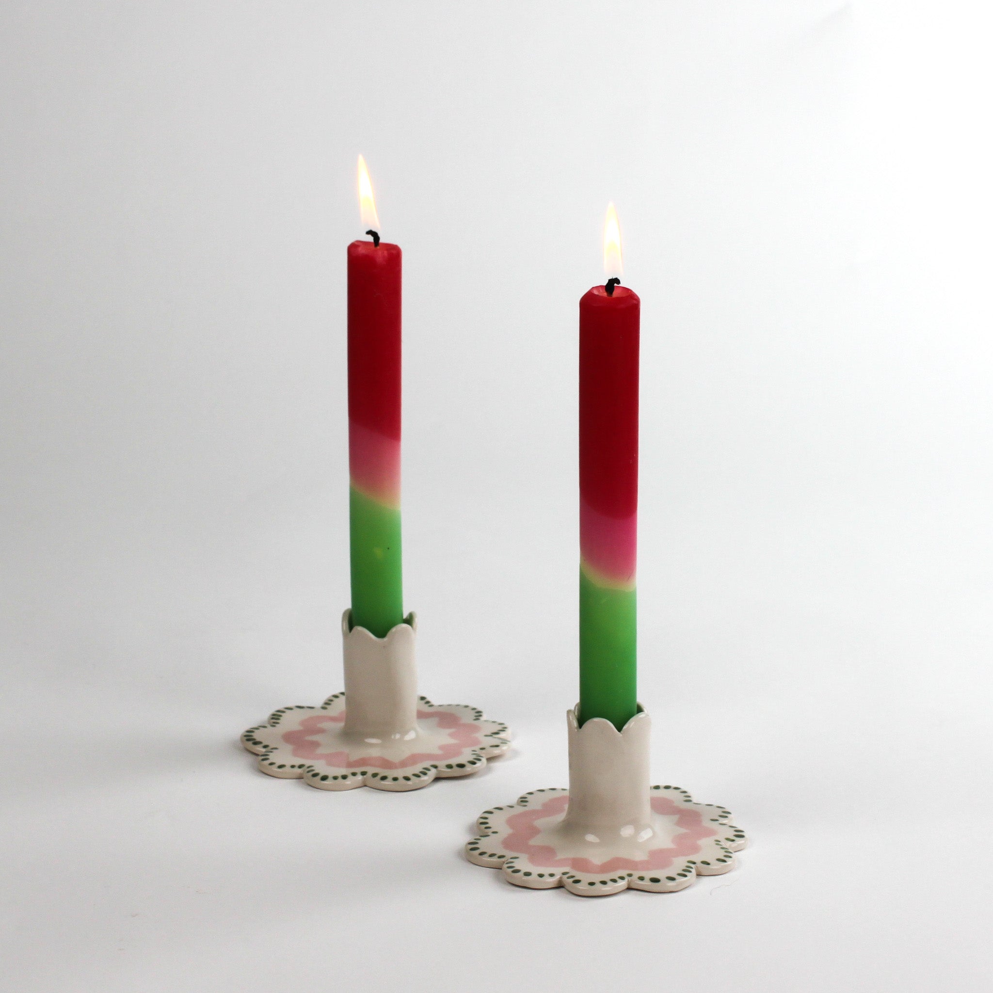 Two lit red-pink-green gradient taper candles in scalloped ceramic candle holders with pink and green detailing, displayed on a white background.