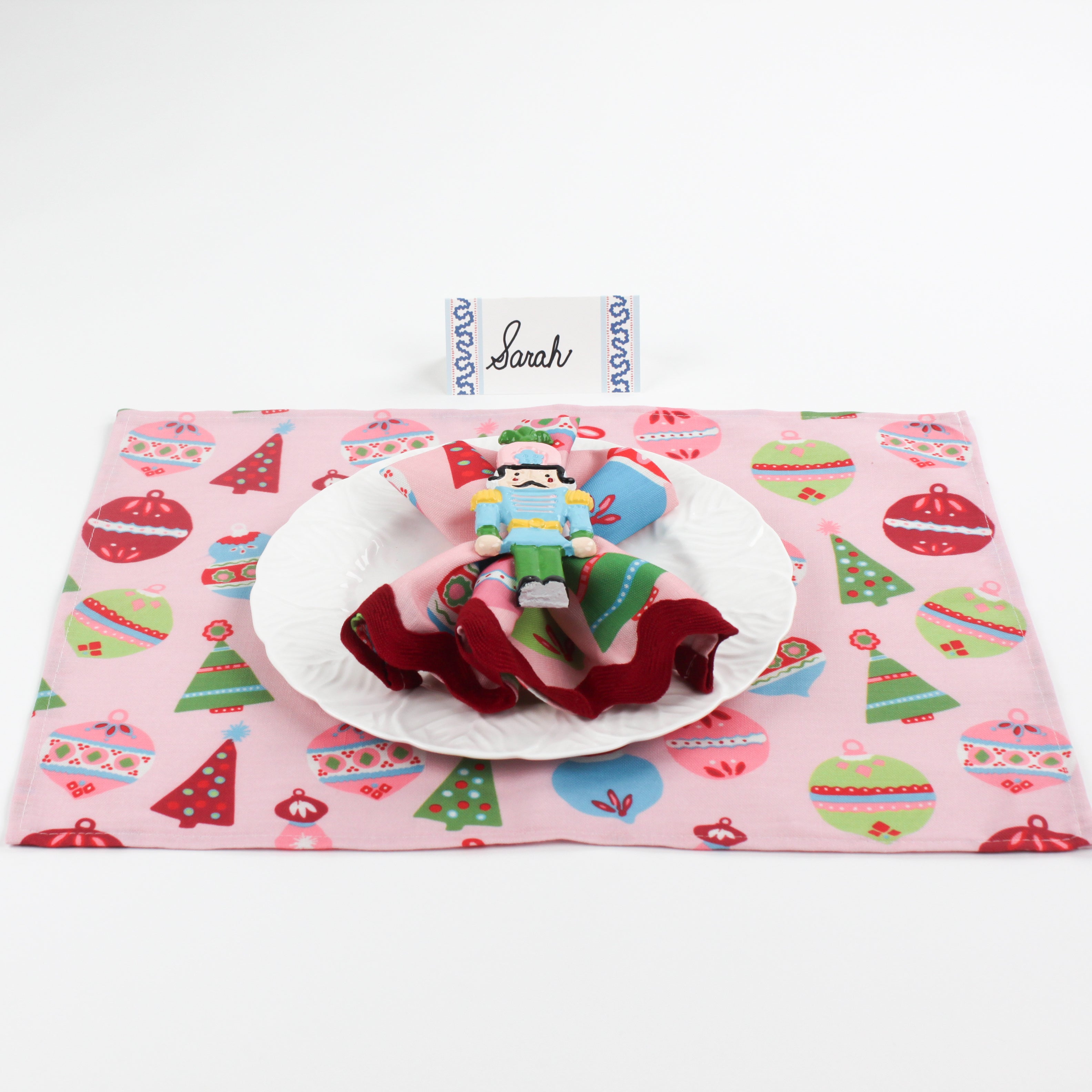 Christmas table setup featuring Harbor Trail Paper Place Card in blue with pink holiday placemat, nutcracker napkin ring, and white plate for festive dining decor.