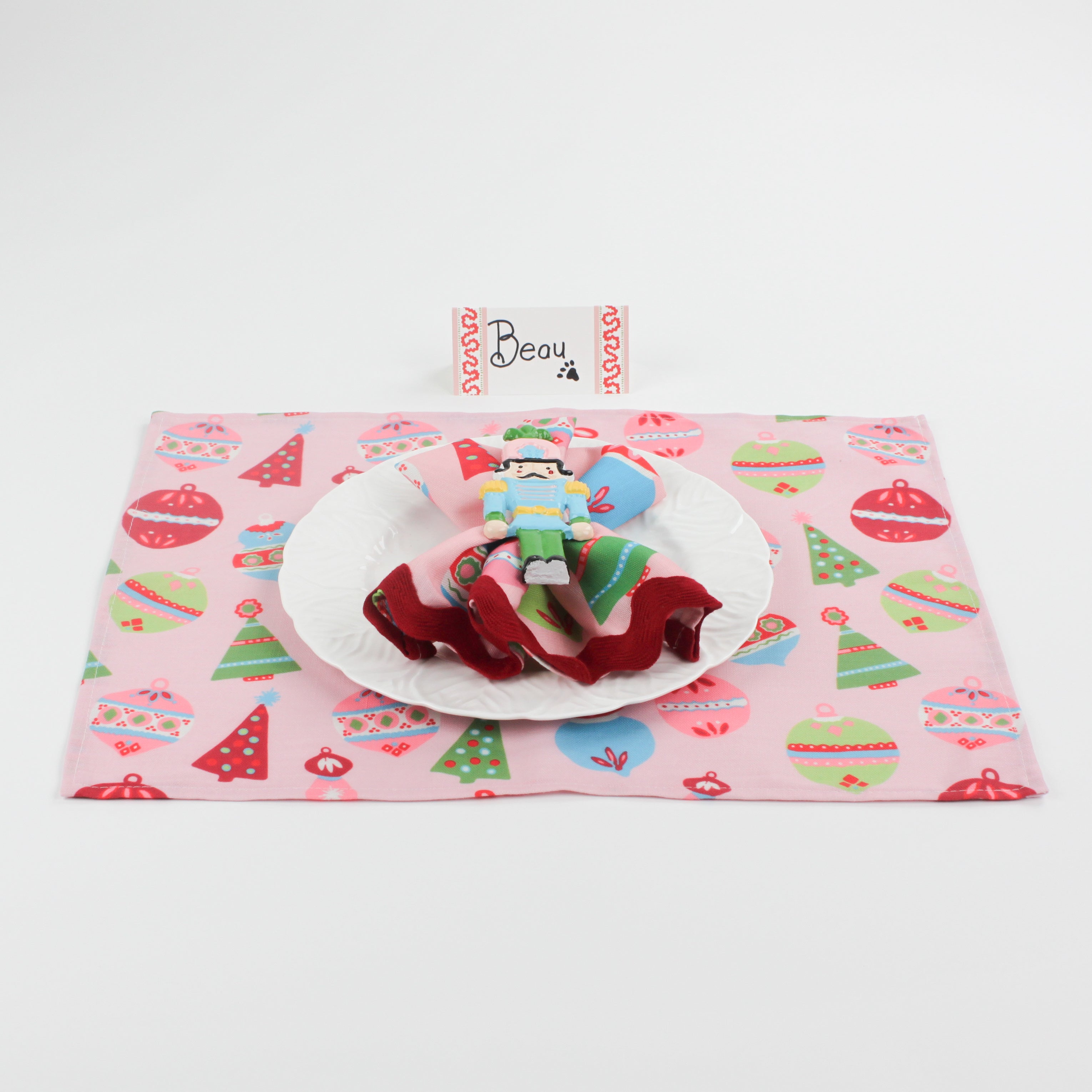 Christmas table setup featuring Harbor Trail Paper Place Card in pink and red, white plate, nutcracker napkin ring, and festive pink placemat with ornaments and trees.
