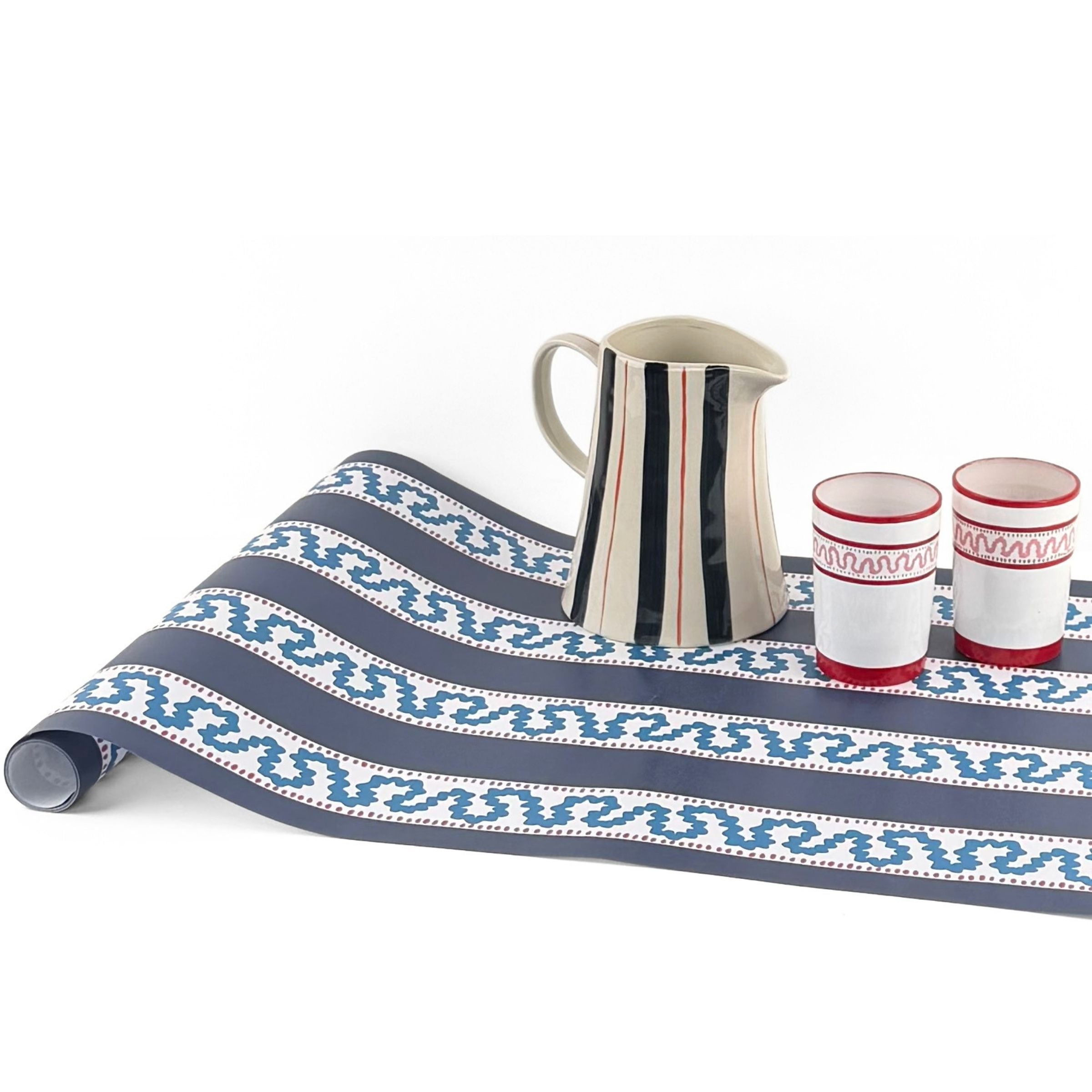 A navy blue and white striped paper table runner with a central light blue wavy stripe on a white background with red glasses and a striped pitcher resting on top. 