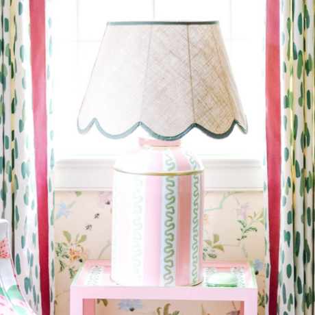 a striped white, pink and green lamp with a scalloped rattan shade with green trim sitting on a pink side table. 