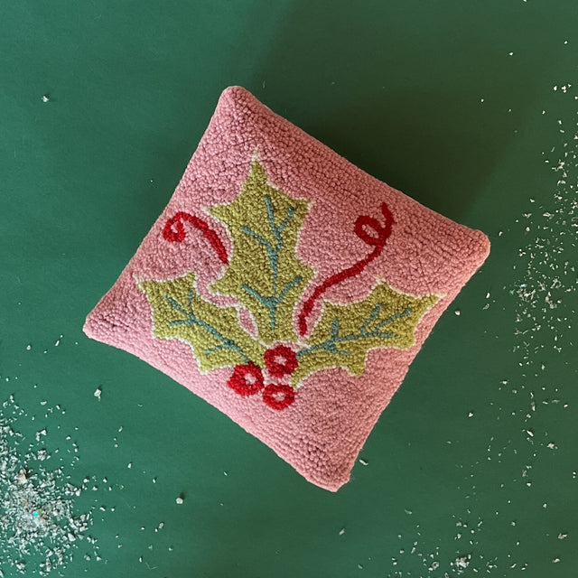 A pink Christmas throw pillow with a large green holly and ribbon motif resting on a green background with a sprinkle of snow