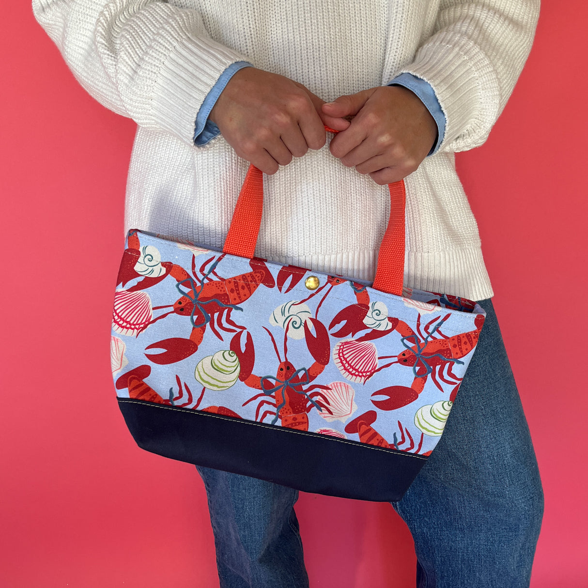 Rock Lobster small canvas tote bag with red straps and blue fabric with a repeating lobster and shell motif held by model