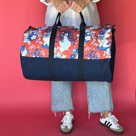 A person holding an Imperial Palace canvas duffle bag with a chinoiserie pattern on the top half and a solid navy blue color on the bottom half.