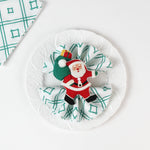 A white dinner plate holding a green-and-white geometric napkin folded in a fan shape, secured with a Santa Claus napkin ring.