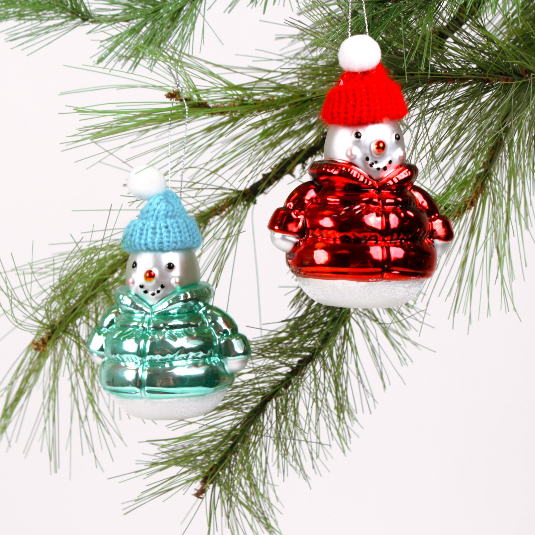 Two glass snowman ornaments, one in a red outfit and blue hat, the other in a green outfit and red hat, hanging on a Christmas tree branch.