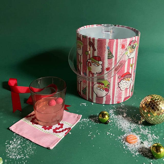 A pink and white stiped fabric with jolly santa motif on a 3 quart acrylic ice bucket surrounded by a cocktail and christmas ornaments