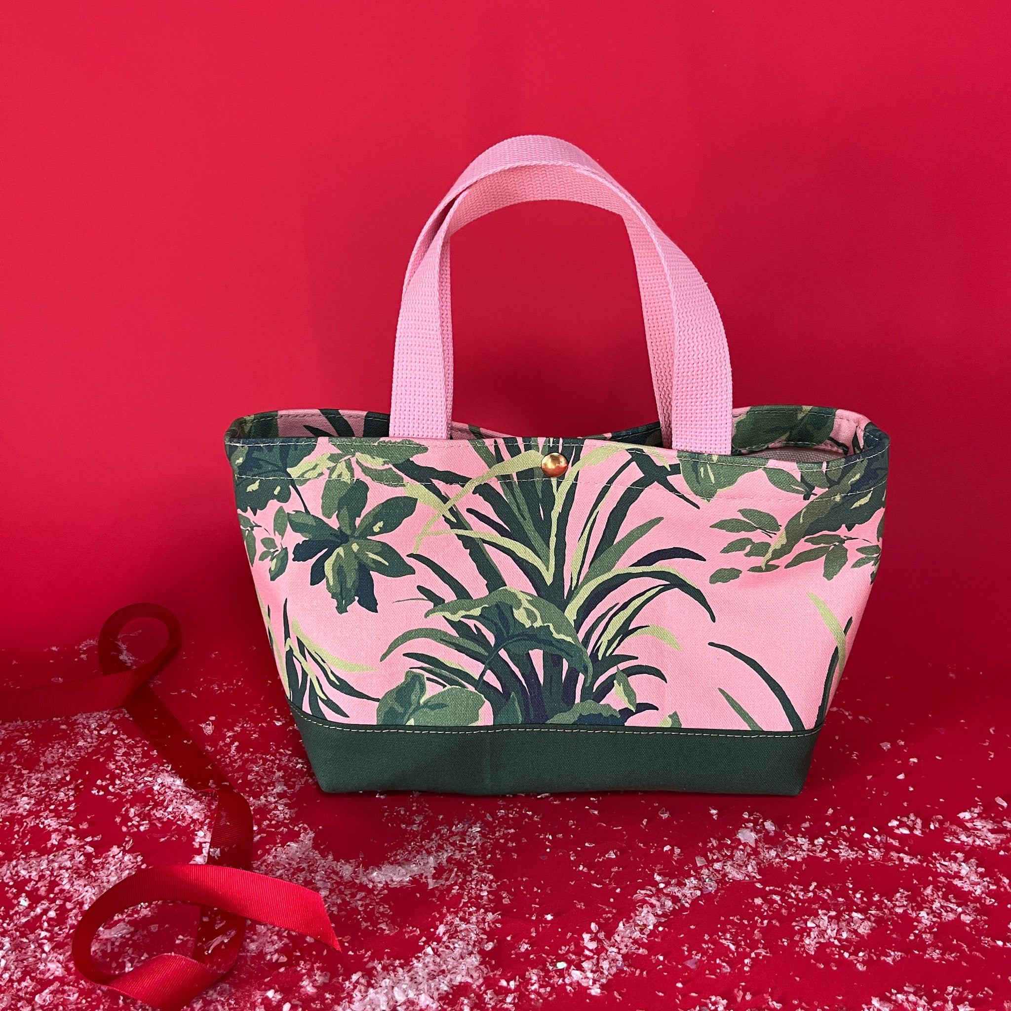 View of the Jungle Road small canvas tote in pink and green on a red background with fake snow around it.