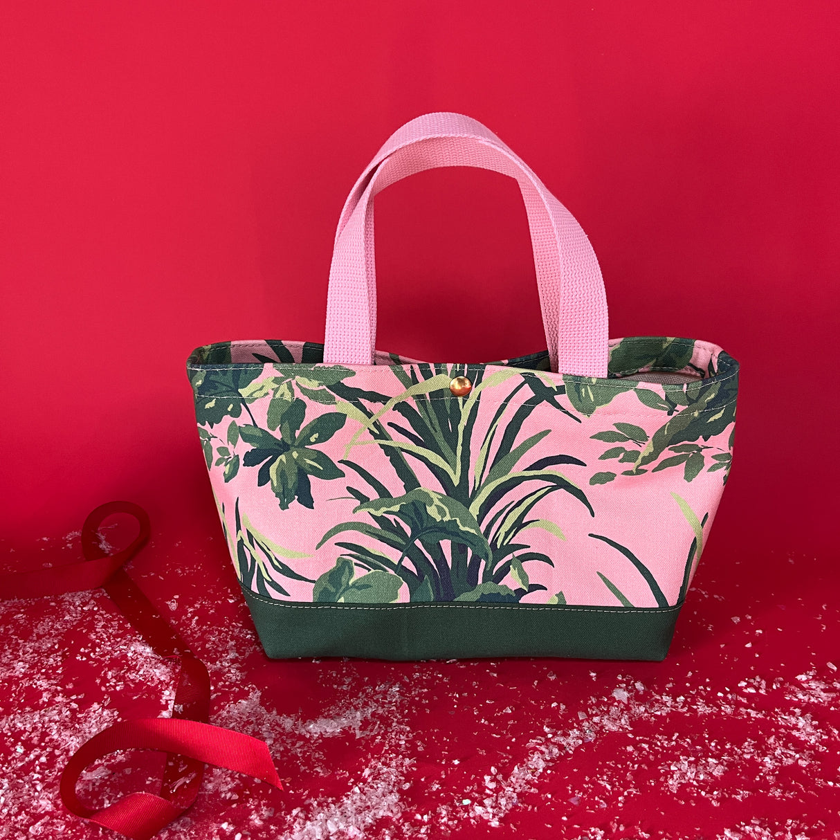 View of the Jungle Road small canvas tote in pink and green on a red background with fake snow around it.