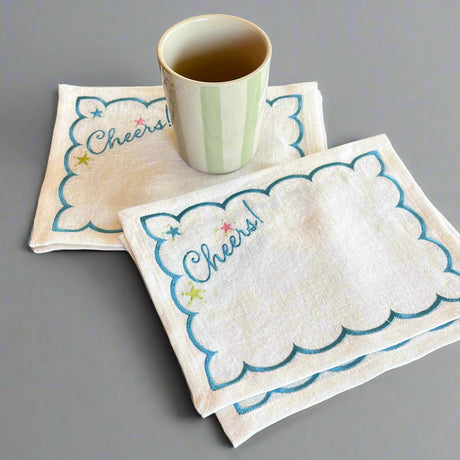Four Cheers! embroidered linen cocktail napkins with a green striped cup on a gray background.