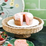 A rattan pedestal bowl with a cake on it, surround by strawberry decor in a green kitchen. 