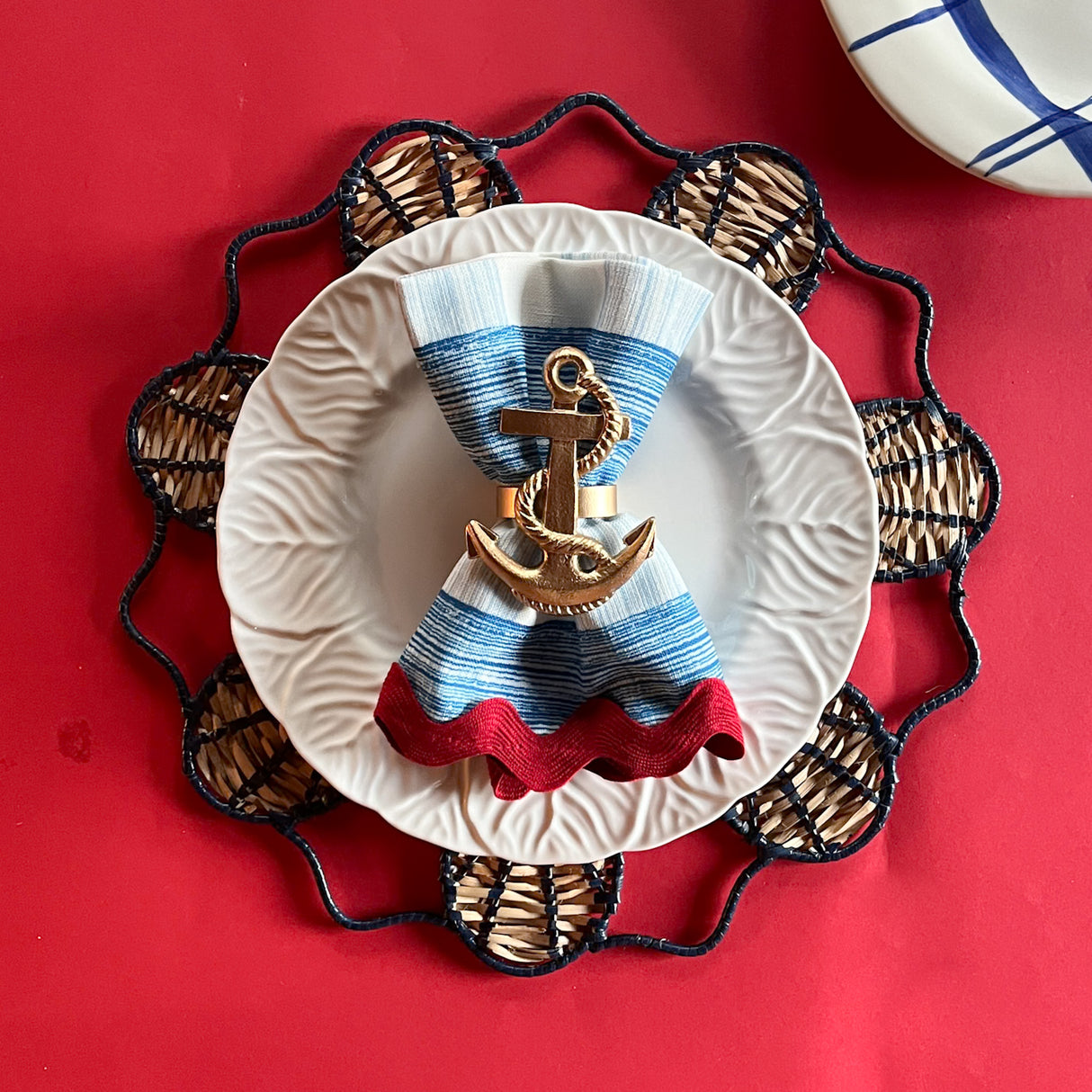Lifestyle shot of the Shell rattan placemat/charger in navy blue with a white plate resting on it, holding a gingham napkin with gold anchor napkin ring.