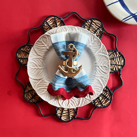 Lifestyle shot of the Shell rattan placemat/charger in navy blue with a white plate resting on it, holding a gingham napkin with gold anchor napkin ring.