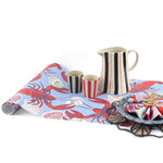 A blue coastal paper table runner with lobster and shell pattern on a white background with a striped pitcher and blue plate resting on top of the runner. 