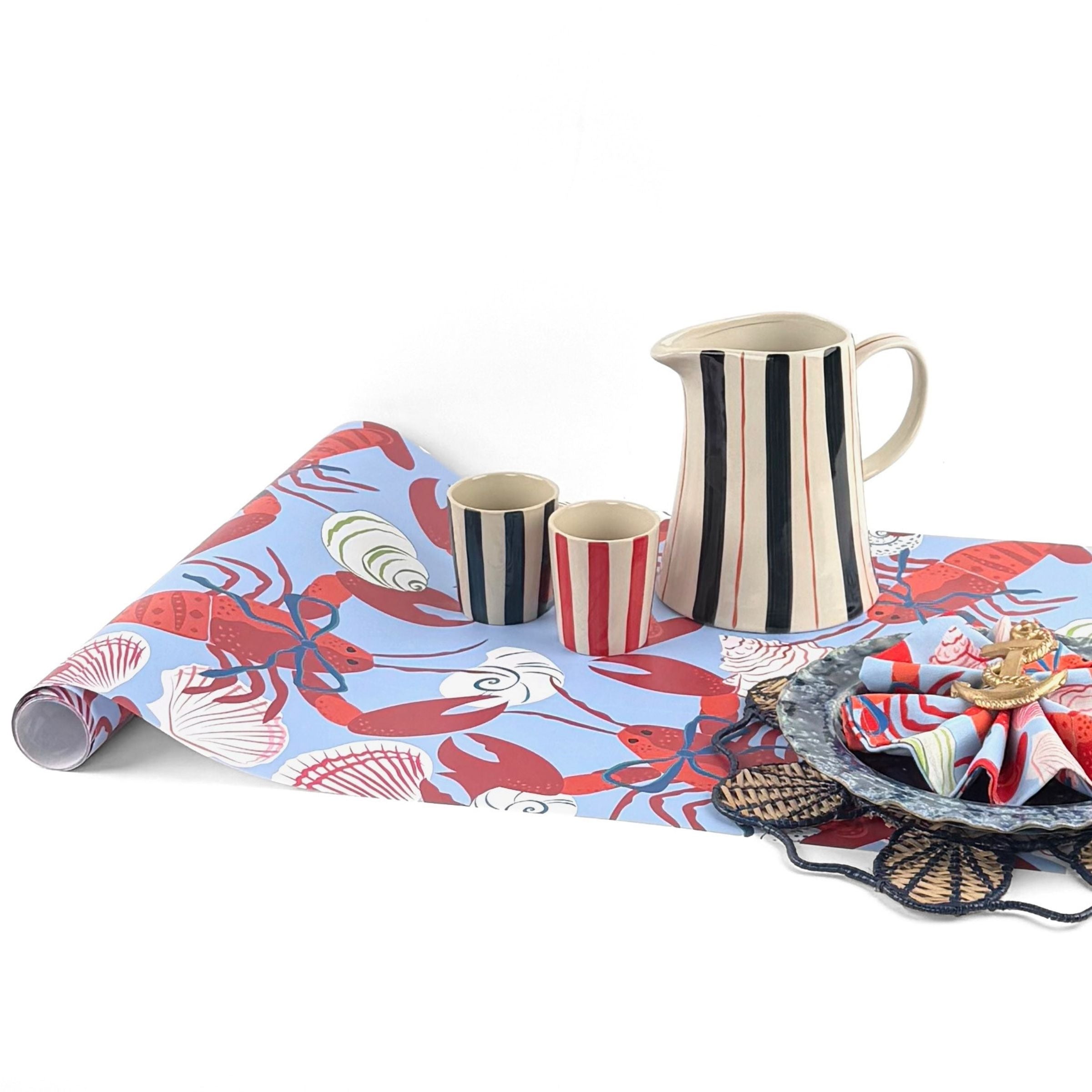 A blue coastal paper table runner with lobster and shell pattern on a white background with a striped pitcher and blue plate resting on top of the runner. 