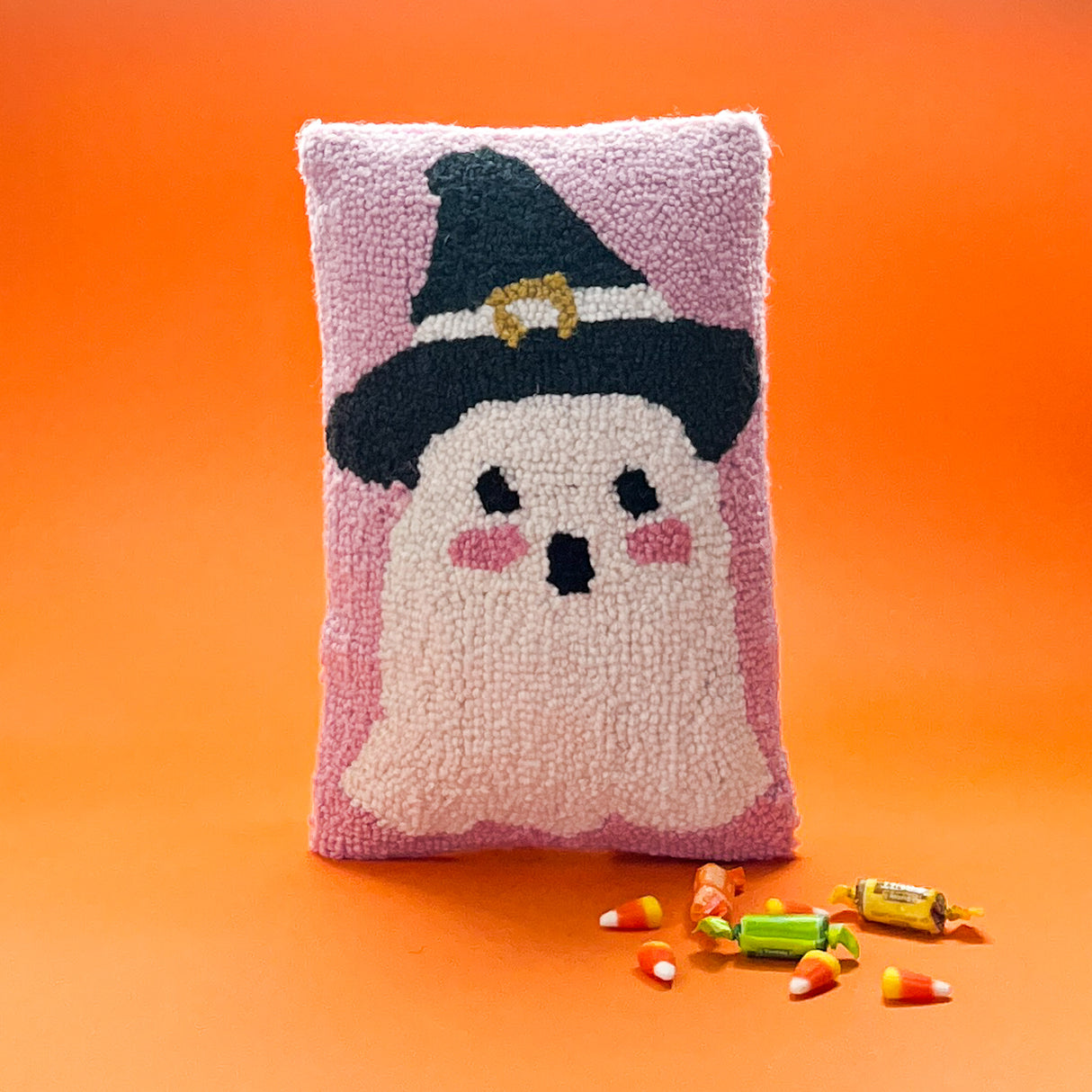 Madcap Cottage Halloween pink Halloween throw pillow with a white ghost with pink cheeks wearing a black witches on an orange background with halloween candy in the foreground.