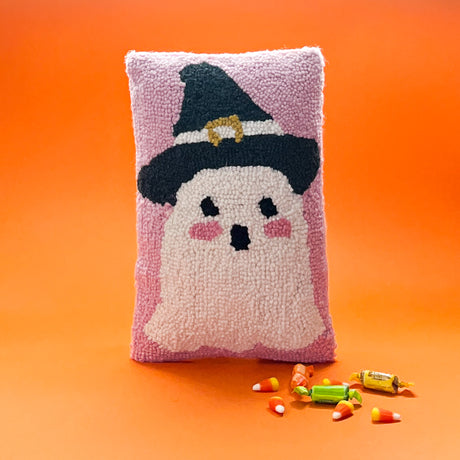 Madcap Cottage Halloween pink Halloween throw pillow with a white ghost with pink cheeks wearing a black witches on an orange background with halloween candy in the foreground.