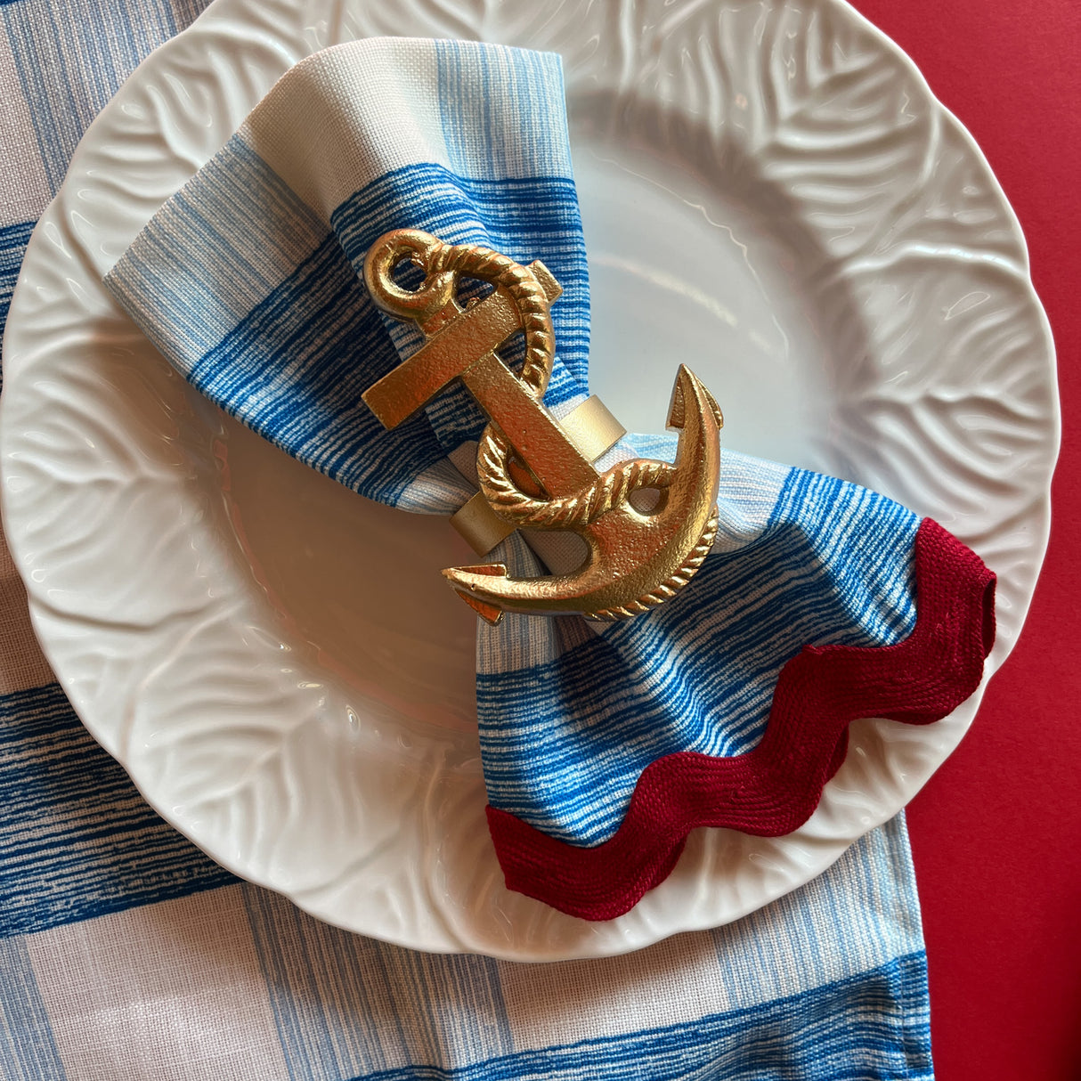 Gin Lane cloth dinner napkin in Swedish Blue with red ric rac trim, folded with a gold anchor napkin ring.