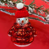 A glass christmas snowman ornament wearing a red hat and coat against a red background