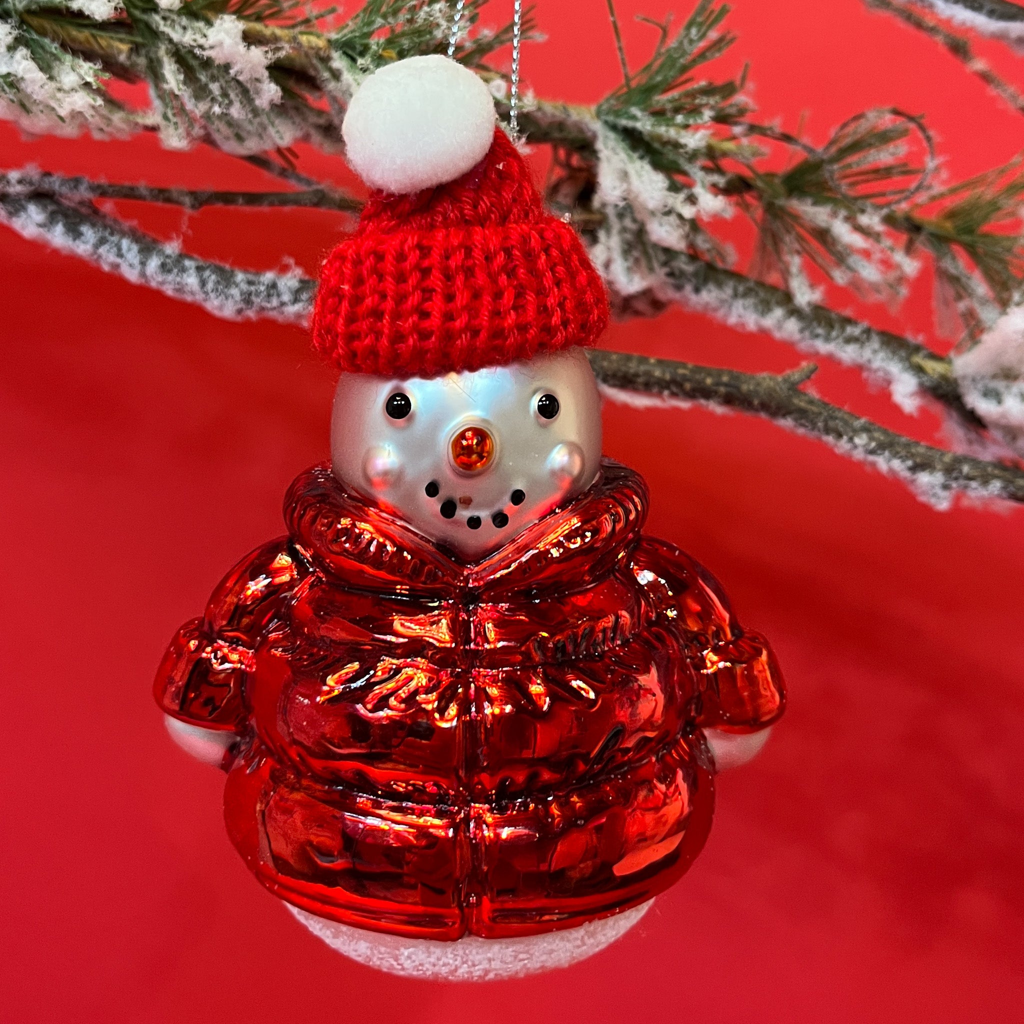 A glass christmas snowman ornament wearing a red hat and coat against a red background