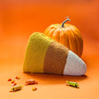 A corn halloween throw pillow shaped like a candy corn with a pumpkin and candy pieces on an orange background.