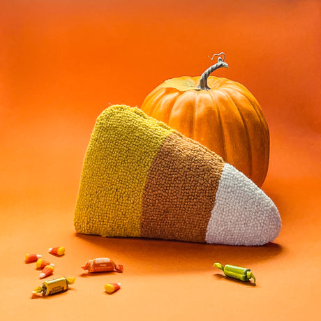A corn halloween throw pillow shaped like a candy corn with a pumpkin and candy pieces on an orange background.