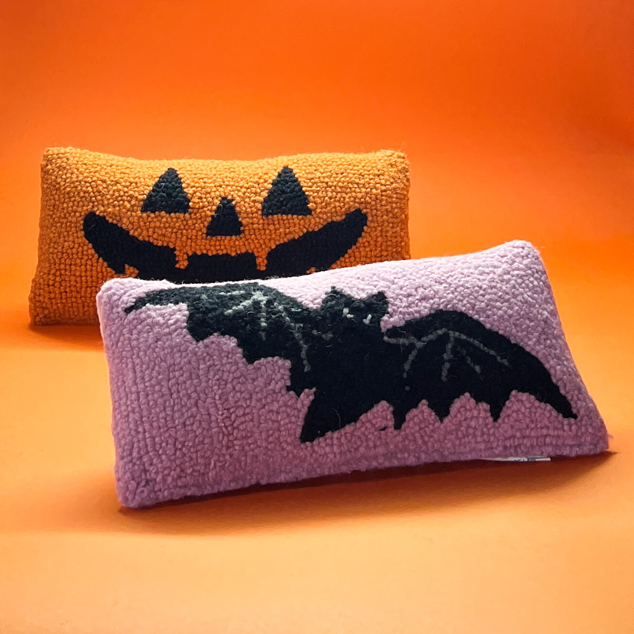 A pink wool lumbar halloween accent pillow with black bat design. another pillow with jack o lantern design