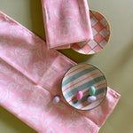 Two floral, shell pink cloth dinner napkins with plaid plates against a green background.