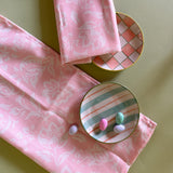 Two floral, shell pink cloth dinner napkins with plaid plates against a green background.
