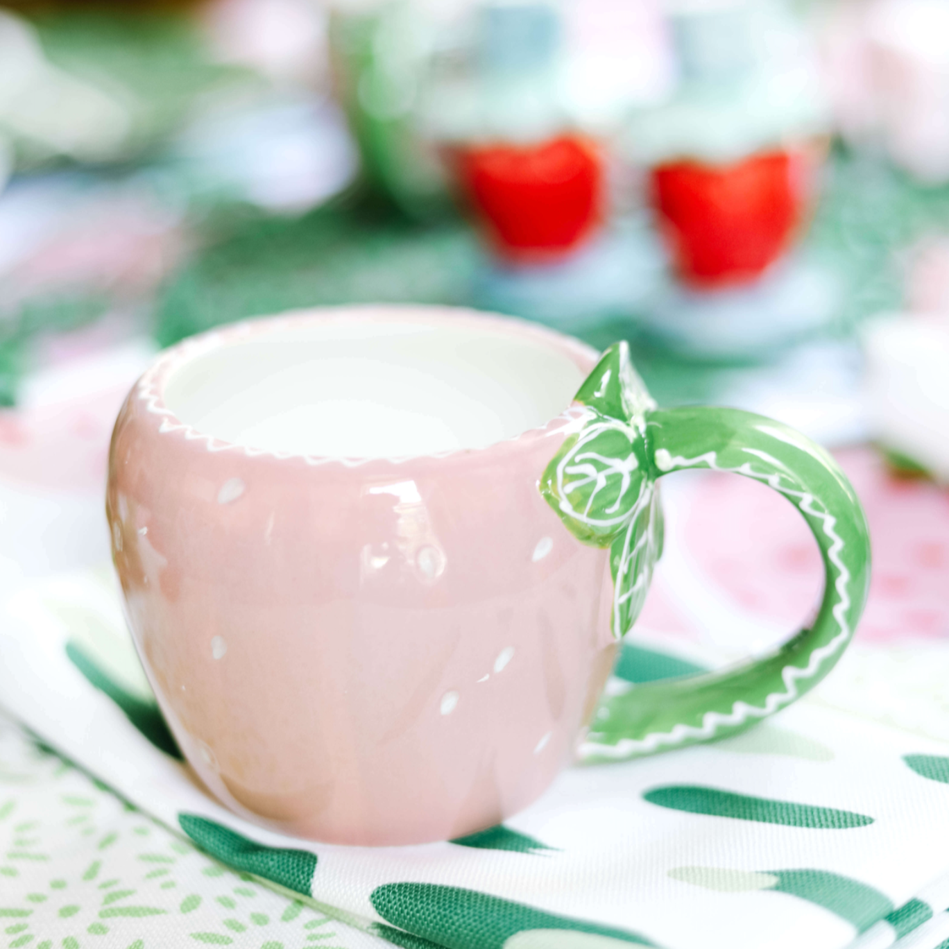 A pink strawberry mug in the shape of a strawberry with a green leafy handle sitting on a green and white napkin