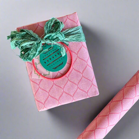 Gift with a pink geometric pattern wrapped with a green bow and ornament embellishment next to the wrapping paper roll