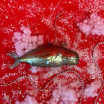 A blue-green whale shaped glass christmas tree ornament against a red, snow covered backdrop