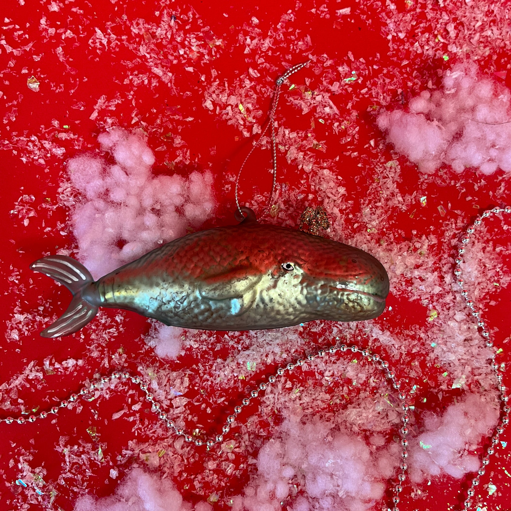 A blue-green whale shaped glass christmas tree ornament against a red, snow covered backdrop