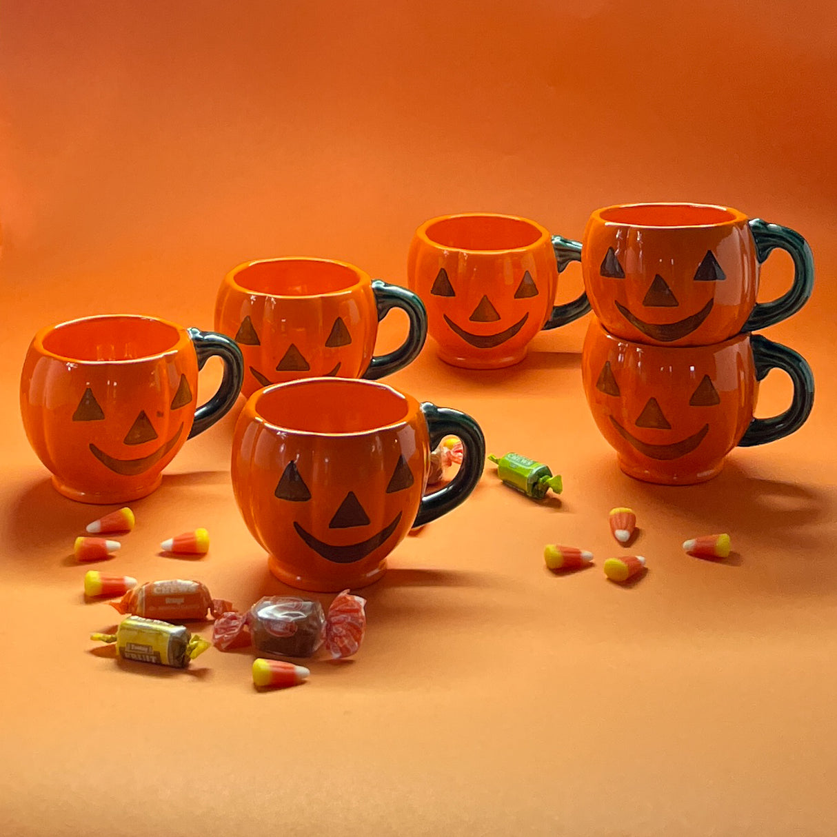 Madcap cottage six jack o'latern halloween coffee mugs on an orange background with halloween candy.