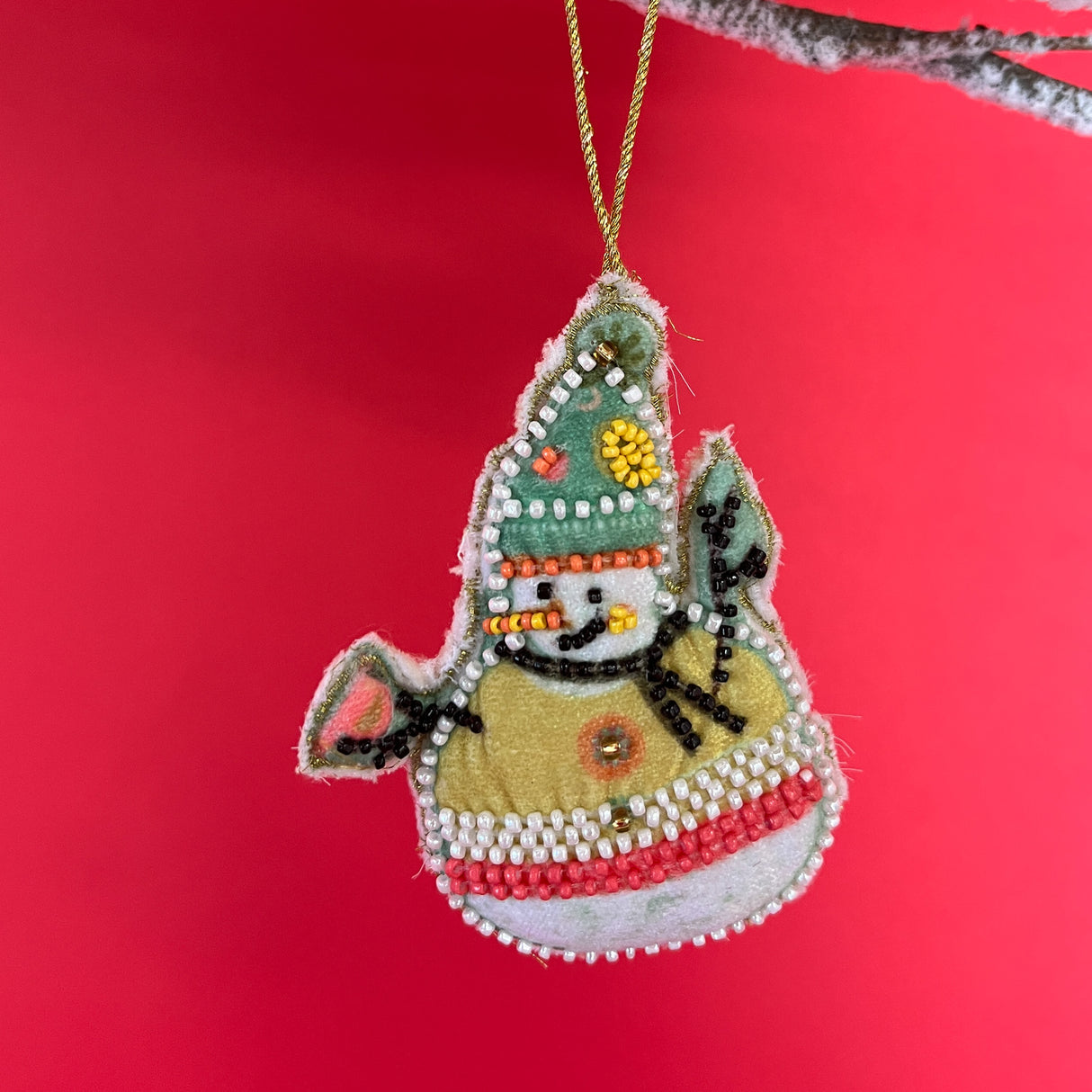 Small snowman ornament with a yellow sweater and small bead details on its hat and sweater on a red background.
