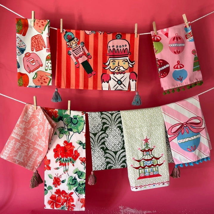 Colorful holiday tea towels hanging on a clothesline against a pink wall, featuring nutcracker, ornaments, pagoda, floral, and chinoiserie designs with tassel accents.