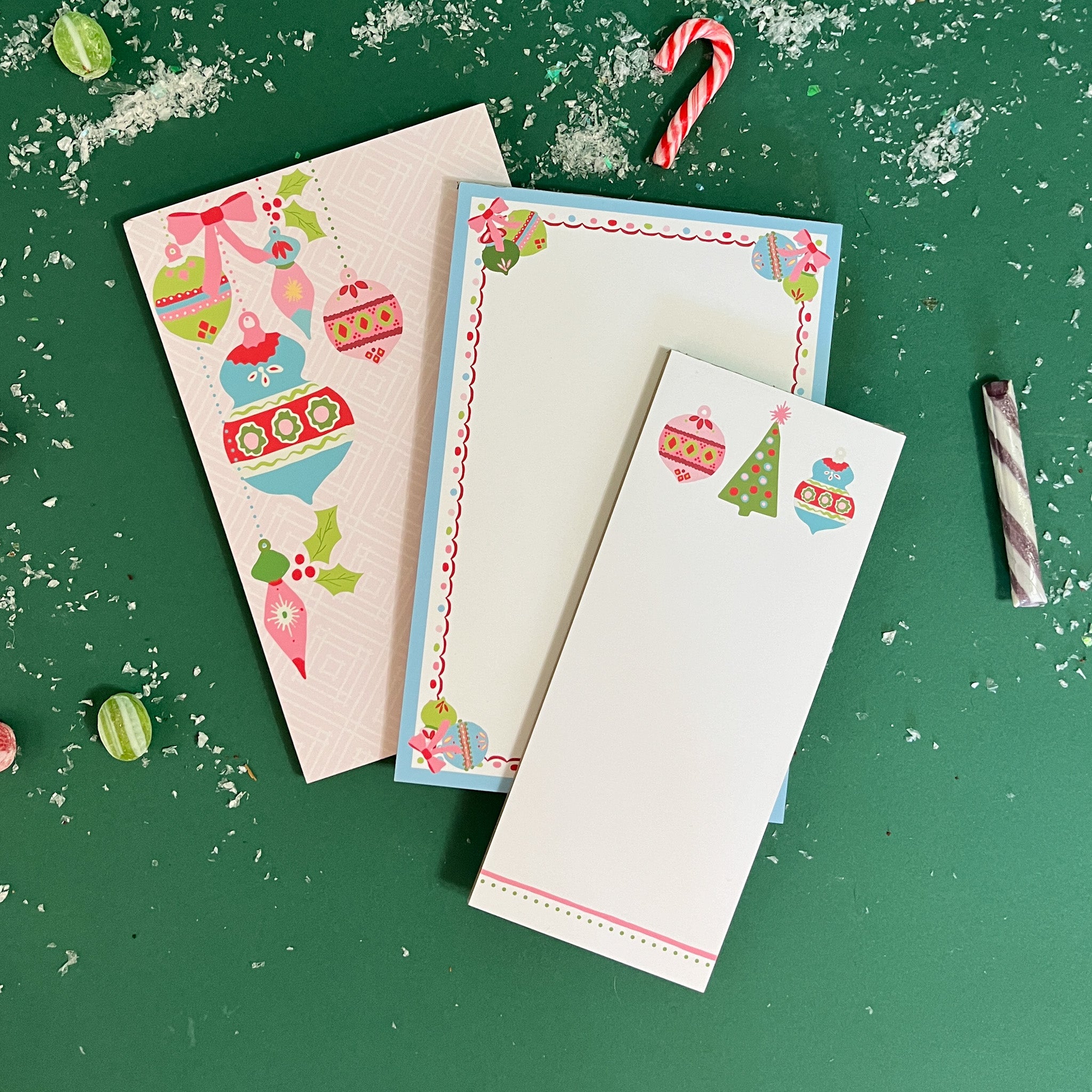 Festive stationery set featuring three pieces: a large sheet with colorful hanging ornaments, a bordered writing pad with playful holiday illustrations, and a slim notepad with ornaments and a Christmas tree, all styled on a green background with candy accents.