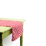 Monserrat Lacquer Red is a no-iron cloth table runner with a geometric pattern, displayed on a green table