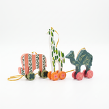 Three hand-painted wooden animal toys with wheels on a clean white background: a pink and green patterned elephant, a white giraffe with green spots, and a teal camel with gold leaf patterns.