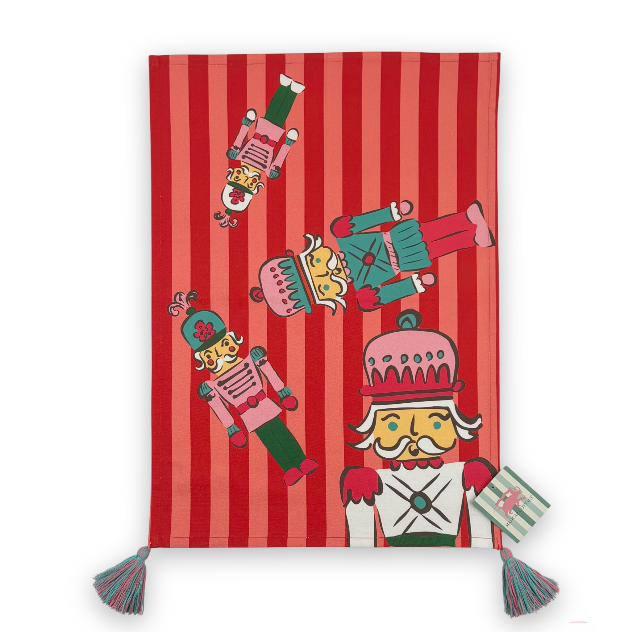 A rectangular red and pink striped tea towel, decorated with cartoon nutcracker illustrations and finished with two tassel ties at the bottom corners and a madcap cottage card on the bottom.