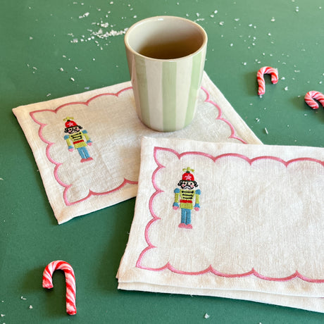 Set of four Nutcracker embroidered linen Christmas cocktail napkins with a striped cup and candy canes on a table.